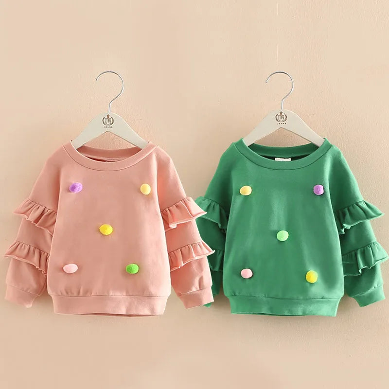 Pink and green girls pom pom jumpers with ruffled sleeves on hangers, suitable for ages 2-10.